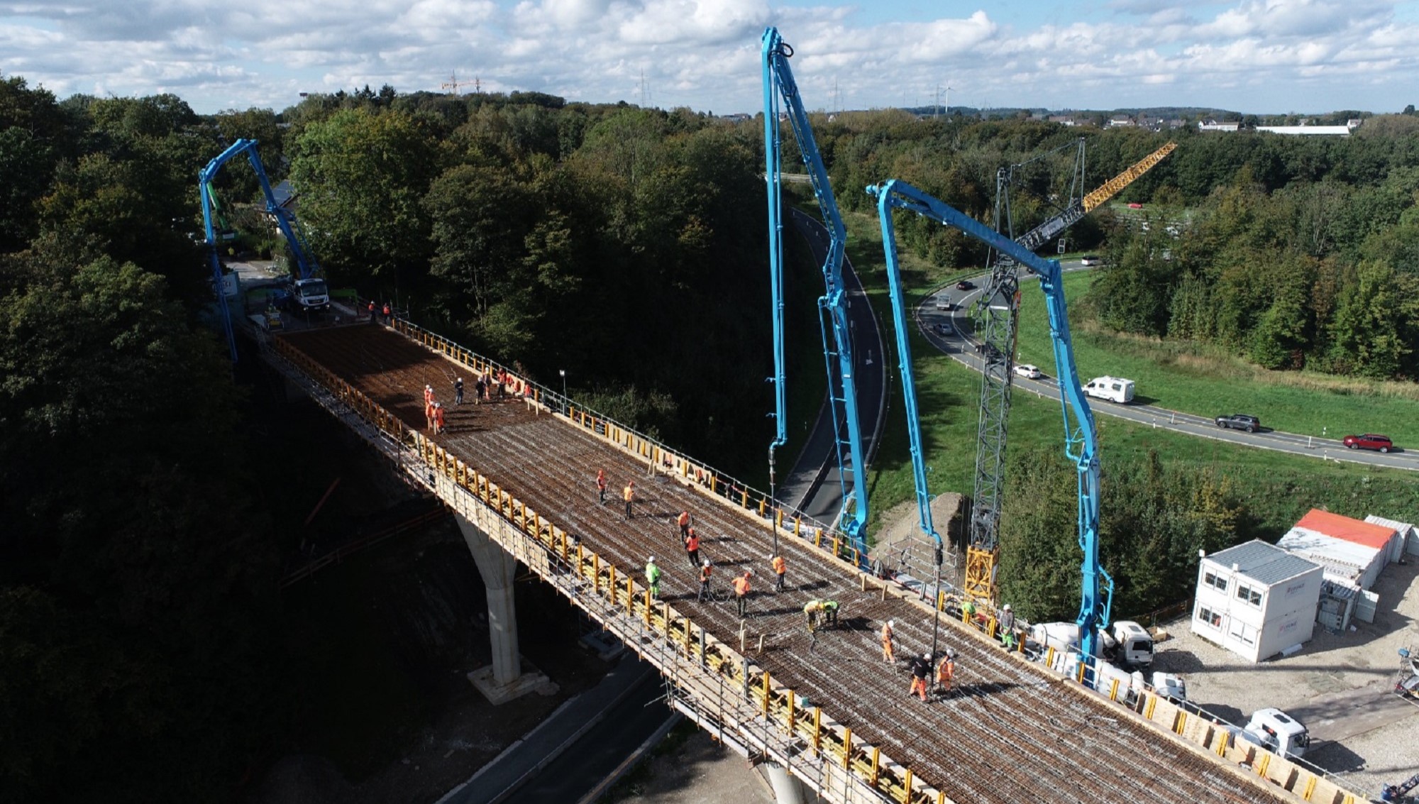 Auf dem Bild ist eine große Brückenbaustelle zu sehen. Mehrere Betonpumpen mit langen Auslegern verteilen frischen Beton, während zahlreiche Arbeiter in Warnwesten die Bewehrung vorbereiten. Die Baustelle liegt inmitten grüner Landschaft mit angrenzender Straße und Baubüros.